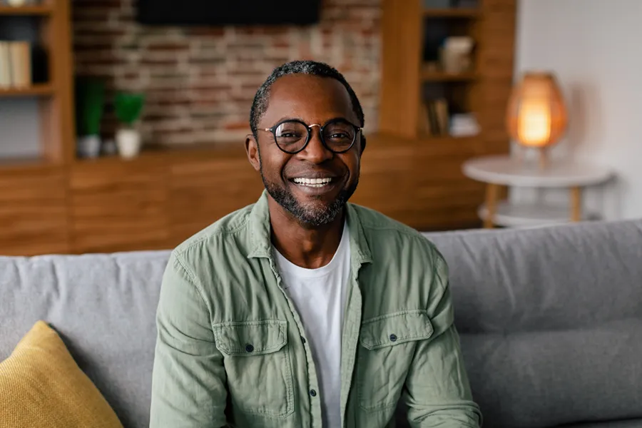 A middle aged black man in a green button up shirt sits on his couch smiling, happy with his treatment for andropause and low testosterone from Blue Skye Health and Wellness in Tampa.