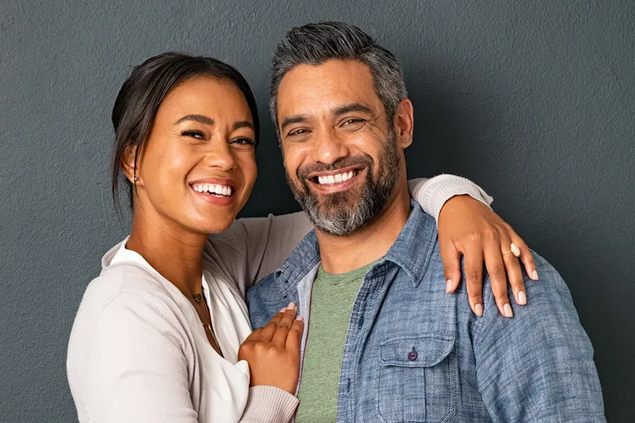 A middle-aged couple with their arms around each other standing against a gray background and smiling, happy with the bioidentical hormone therapy they received from Blue Skye Health and Wellness in Tampa.