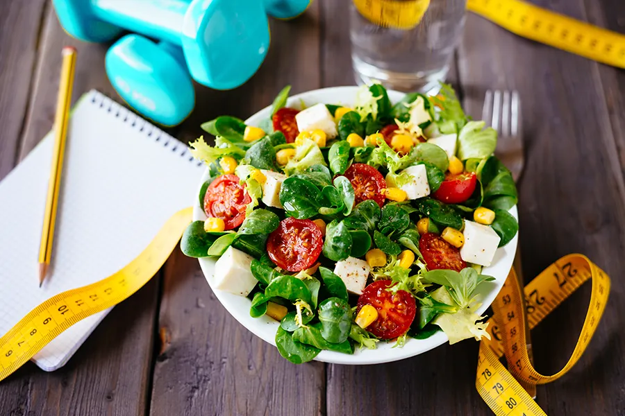 A tableau on a wooden table of a nutritious salad, barbells, and a measuring tape. Get info about the Blue Skye Body Program from Blue Skye Health and Wellness in Tampa.