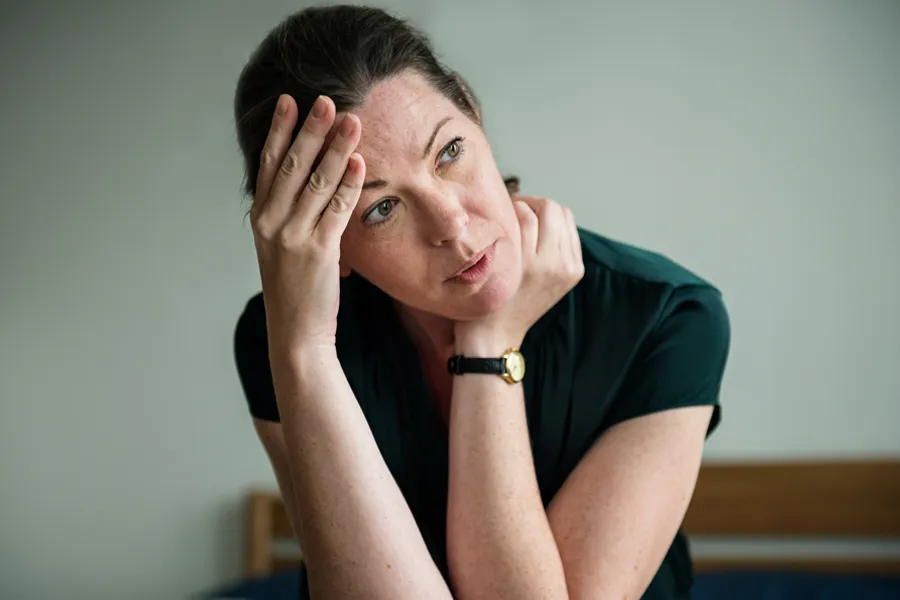 A woman in a dark blue dress sits on her bed holding her head in her hands with an exhausted expression. Get help for chronic fatigue syndrome from Blue Skye Health and Wellness in [AERA].