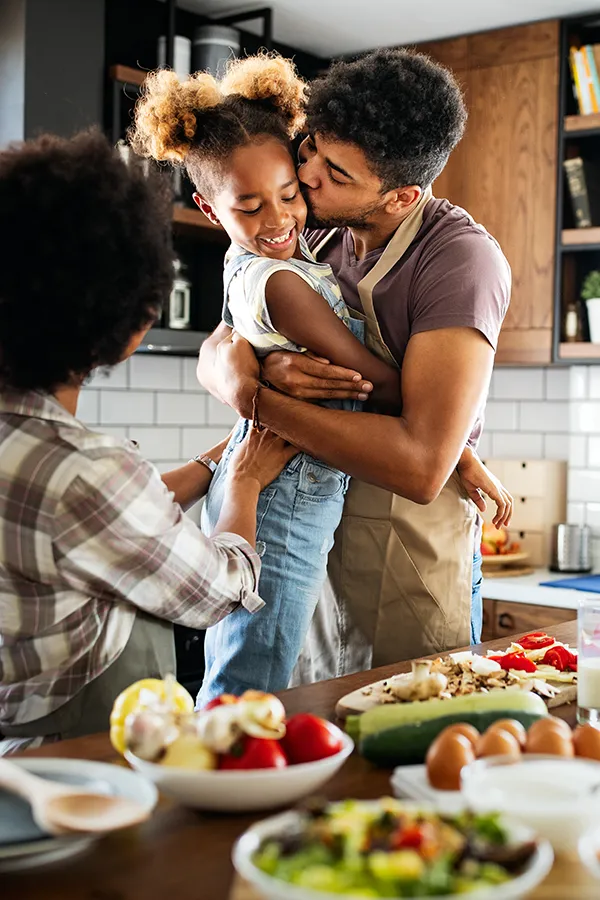 A man holds his daughter and kisses her cheek as the family makes a healthy dinner in the kitchen. Get help for digestive issues and gut health from Blue Skye Health and Wellness in Tampa.