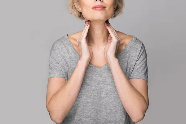 An older blonde woman in a gray t-shirt holds her hands to her neck checking for signs of thyroid problems. Get help for Hashimoto's Thyroiditis from Blue Skye Health and Wellness in Tampa.