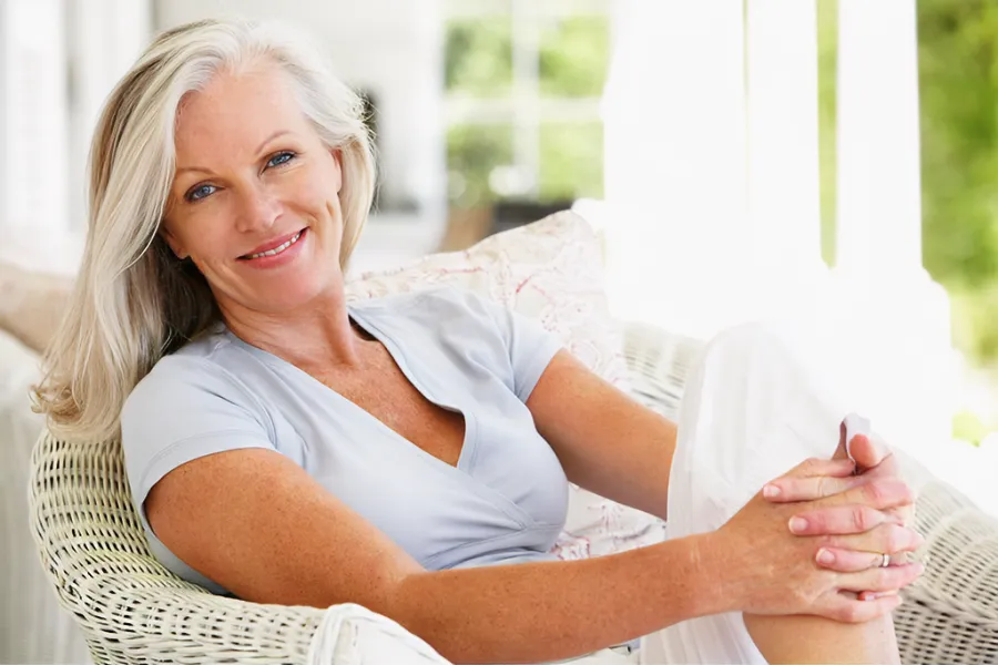 A smiling mature woman sits on a wicker chair on her covered porch. Get hormones for menopause from Blue Skye Health and Wellness in Tampa.
