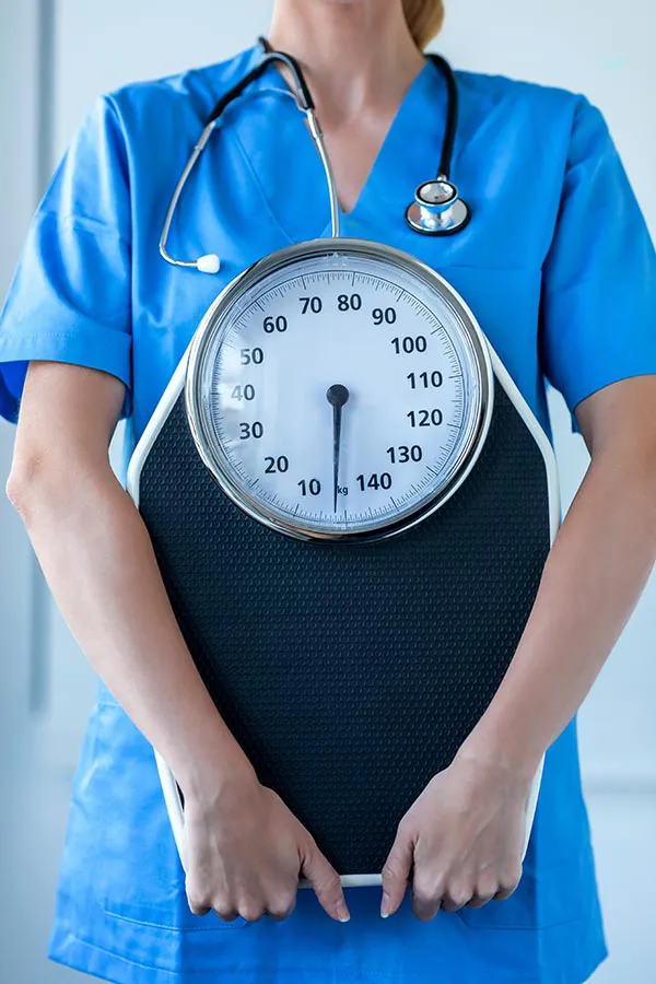 A nurse in blue scrubs with a stethoscope hanging around her neck holds a scale, representing weight loss treatments from Blue Skye Health and Wellness in Tampa.