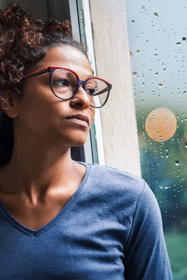 A black woman with with a somber expression and glasses and a gray shirt stares out the window on a rainy day. Get treatment for Premenstrual Dysphoric Disorder from Blue Skye Health and Wellness in Tampa.