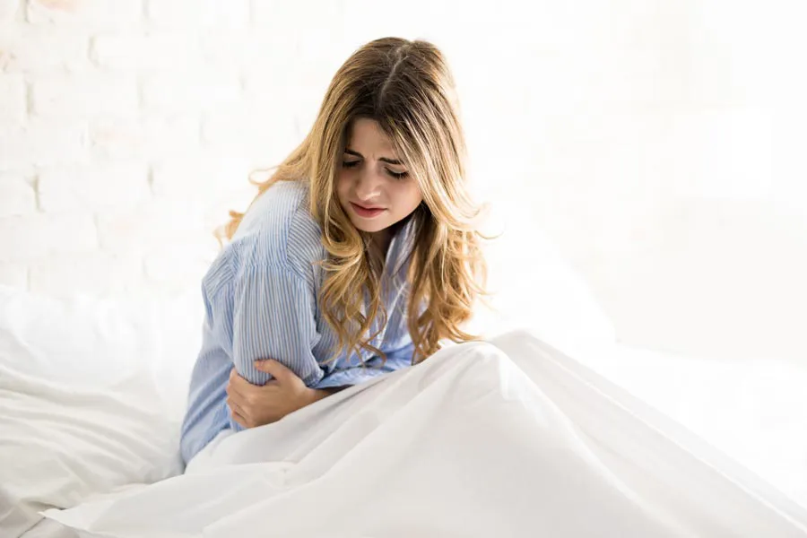 A woman with long blonde hair sits in her bed holding her stomach in pain, considering getting care for PMS from Blue Skye Health and Wellness in Tampa.