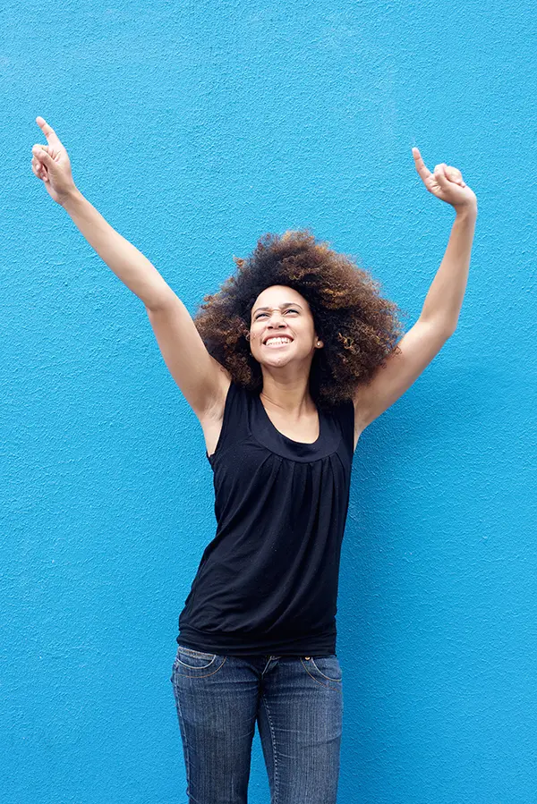 A woman in a dark blue tank top standing in front of a bright blue wall, raising her arms in celebration of relief from PMS from Blue Skye Health and Wellness in Tampa.
