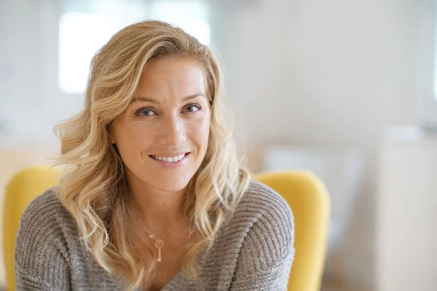 A blonde woman wearing a a gray sweater sits inside in a yellow chair, smiling and happy with her perimenopause treatment from Blue Skye Health and Wellness in Tampa.