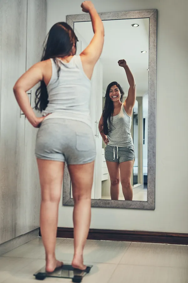 A woman in workout clothes stands on a scale looking in the mirror and pumping her hands in celebration of her successful Tirzepatide treatment from Blue Skye Health and Wellness in Tampa.